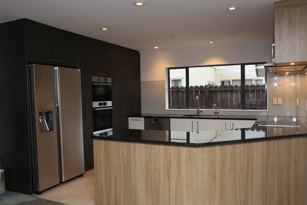 Inspiration for a large contemporary u-shaped open plan kitchen in Auckland with an undermount sink, flat-panel cabinets, medium wood cabinets, granite benchtops, grey splashback, glass sheet splashback, stainless steel appliances, porcelain floors, a peninsula, beige floor and black benchtop.