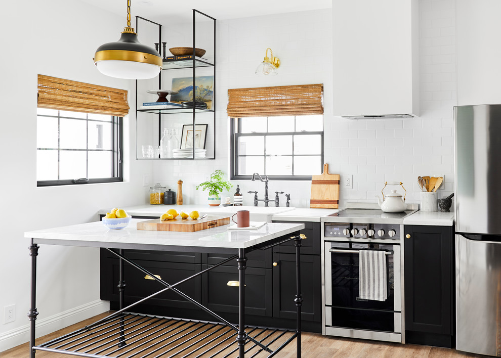 Small mediterranean single-wall open plan kitchen in Los Angeles with a farmhouse sink, shaker cabinets, black cabinets, quartz benchtops, white splashback, stainless steel appliances, light hardwood floors, with island, white benchtop and subway tile splashback.