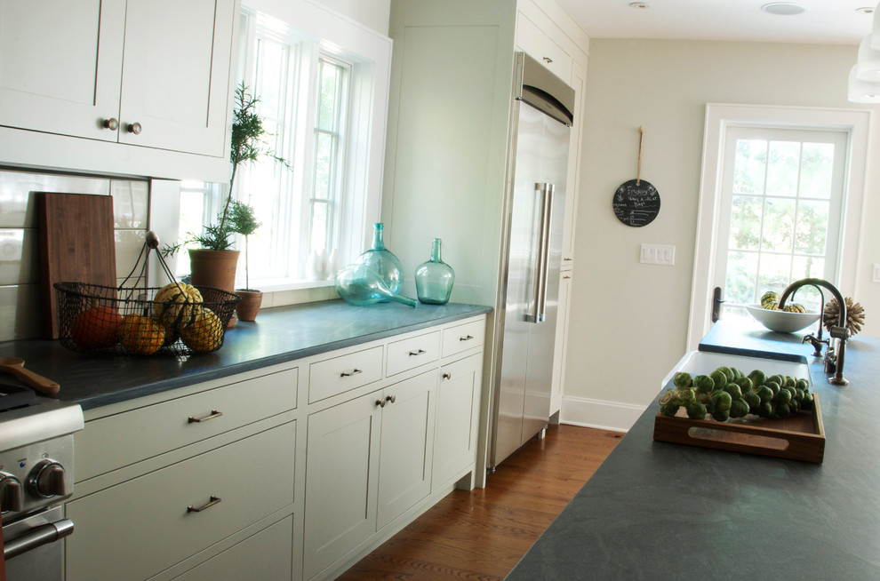 Transitional l-shaped kitchen photo in New York with a farmhouse sink, shaker cabinets, gray cabinets, limestone countertops, gray backsplash, subway tile backsplash and stainless steel appliances