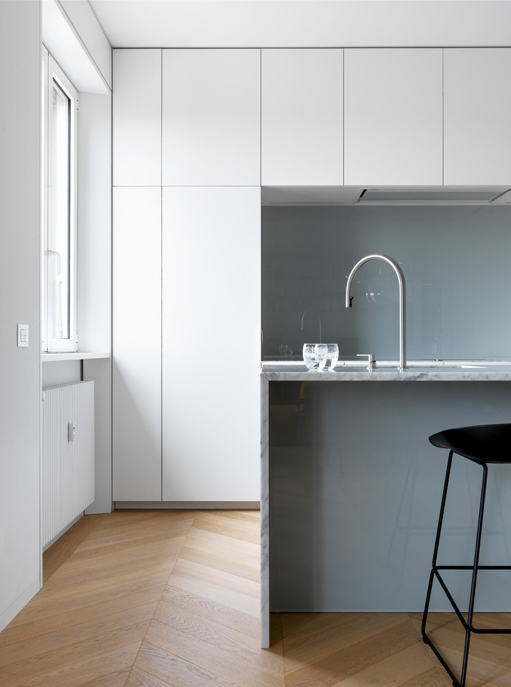 Photo of a large contemporary single-wall eat-in kitchen in Milan with an undermount sink, flat-panel cabinets, white cabinets, marble benchtops, grey splashback, glass sheet splashback, panelled appliances, medium hardwood floors, with island, beige floor and white benchtop.