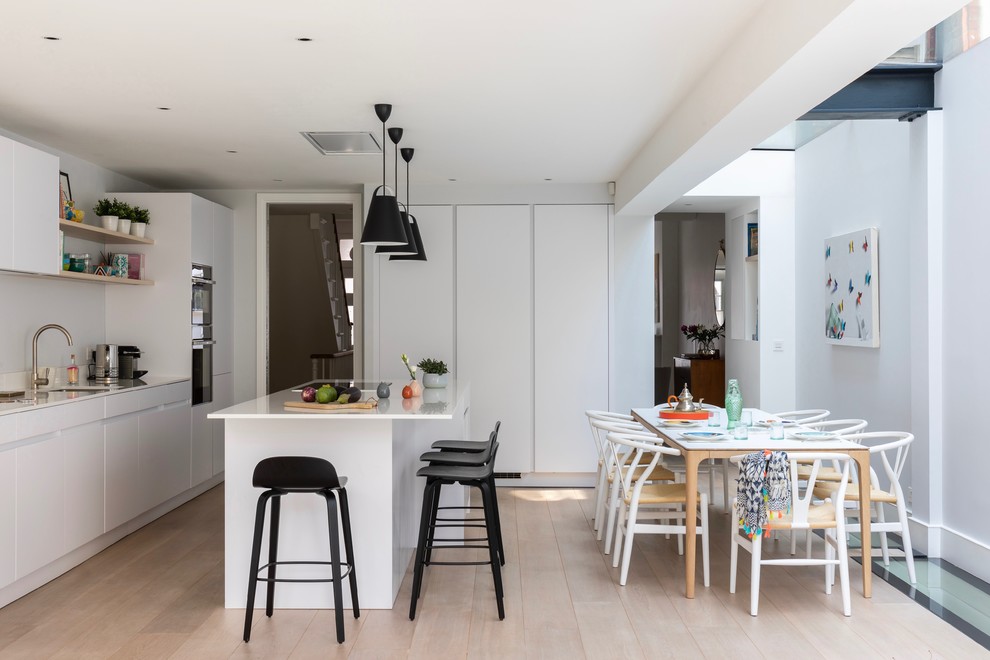 Design ideas for a large contemporary galley eat-in kitchen in London with an integrated sink, flat-panel cabinets, white cabinets, limestone benchtops, white splashback, limestone splashback, black appliances, light hardwood floors, with island, beige floor and white benchtop.