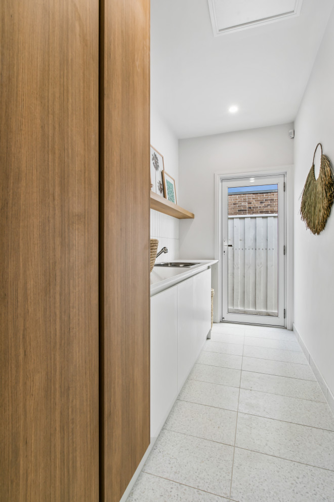 Photo of a mid-sized contemporary single-wall dedicated laundry room in Melbourne with a single-bowl sink, white cabinets, white splashback, subway tile splashback, white walls, porcelain floors, a concealed washer and dryer, white floor and white benchtop.