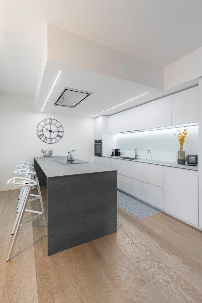 Inspiration for a mid-sized modern single-wall open plan kitchen in Other with a drop-in sink, flat-panel cabinets, white cabinets, quartz benchtops, white splashback, glass sheet splashback, stainless steel appliances, painted wood floors, with island and grey benchtop.