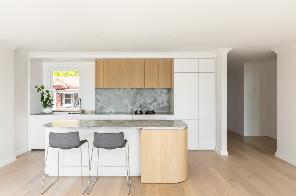 Mid-sized contemporary galley open plan kitchen in Sydney with an integrated sink, flat-panel cabinets, light wood cabinets, stainless steel benchtops, grey splashback, stone slab splashback, panelled appliances, light hardwood floors, with island and grey benchtop.
