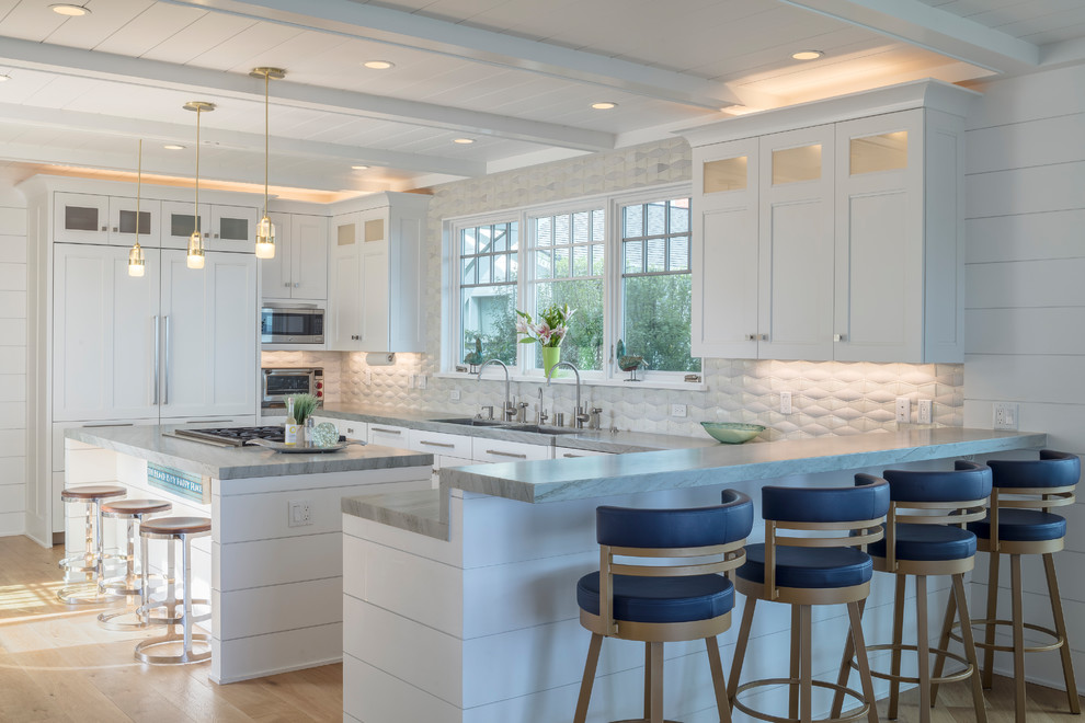 This is an example of a beach style u-shaped kitchen in Sacramento with a double-bowl sink, shaker cabinets, white cabinets, marble benchtops, white splashback, white appliances, light hardwood floors, with island and grey benchtop.