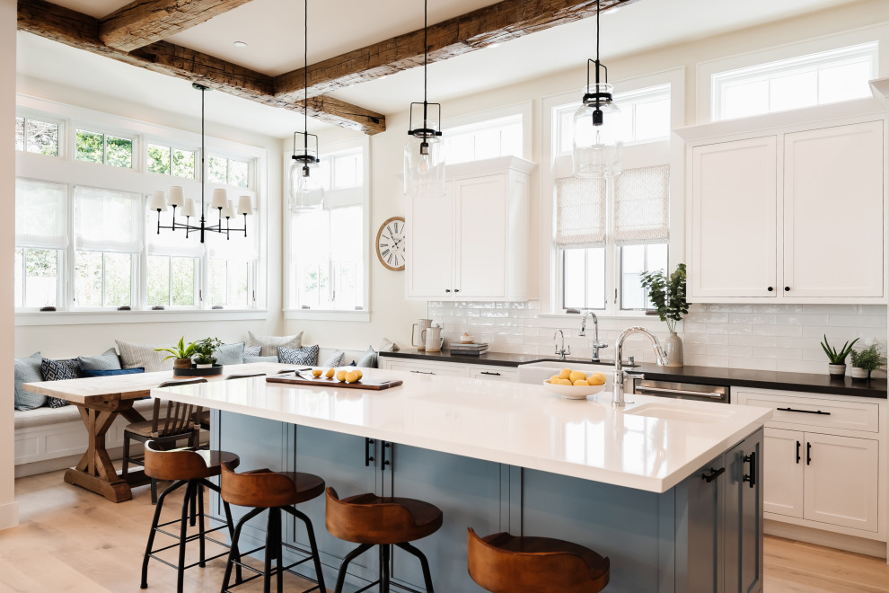 Inspiration for a large farmhouse u-shaped open plan kitchen in San Diego with a belfast sink, shaker cabinets, white cabinets, engineered stone countertops, white splashback, porcelain splashback, stainless steel appliances, light hardwood flooring, an island, brown floors, black worktops and exposed beams.