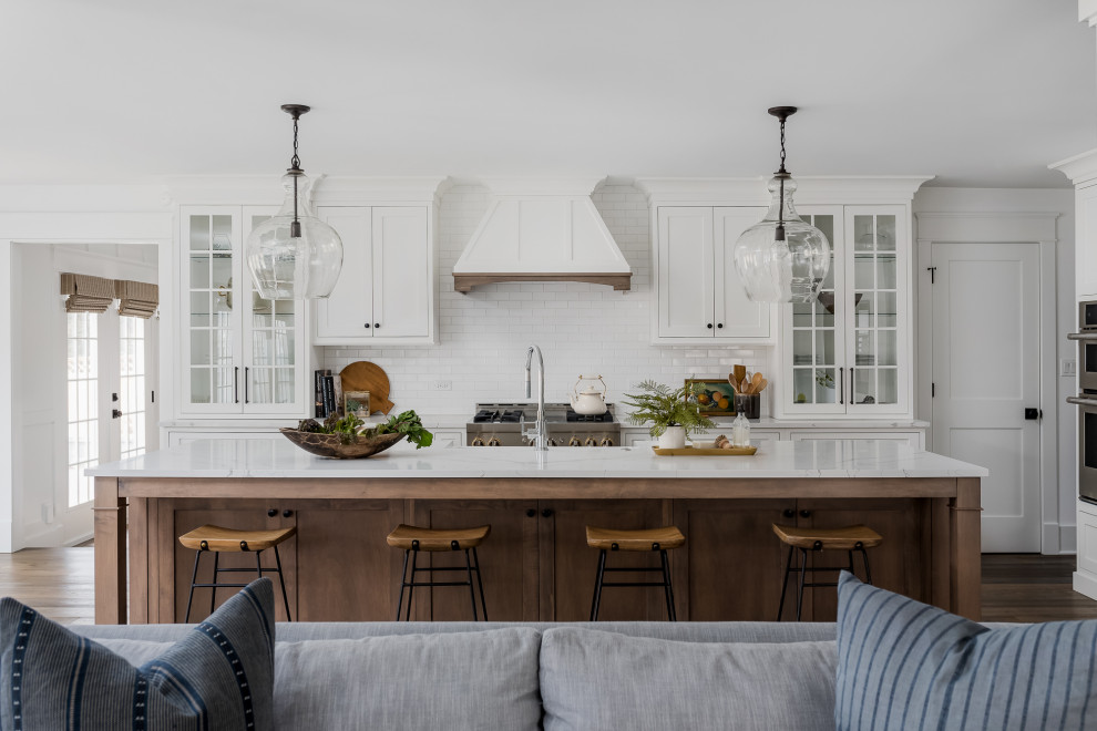 Design ideas for a large country l-shaped open plan kitchen in Chicago with a farmhouse sink, shaker cabinets, white cabinets, quartz benchtops, white splashback, stainless steel appliances, medium hardwood floors, with island, brown floor, white benchtop and subway tile splashback.
