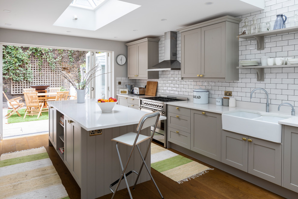 This is an example of a transitional kitchen in London with a farmhouse sink, grey cabinets, white splashback, subway tile splashback, stainless steel appliances, with island, white benchtop, shaker cabinets and dark hardwood floors.