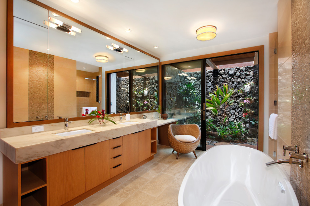 Photo of a large tropical master bathroom in Hawaii with flat-panel cabinets, light wood cabinets, a freestanding tub, a one-piece toilet, beige tile, stone tile, white walls, travertine floors, an undermount sink, granite benchtops, beige floor, beige benchtops, a double vanity and a built-in vanity.