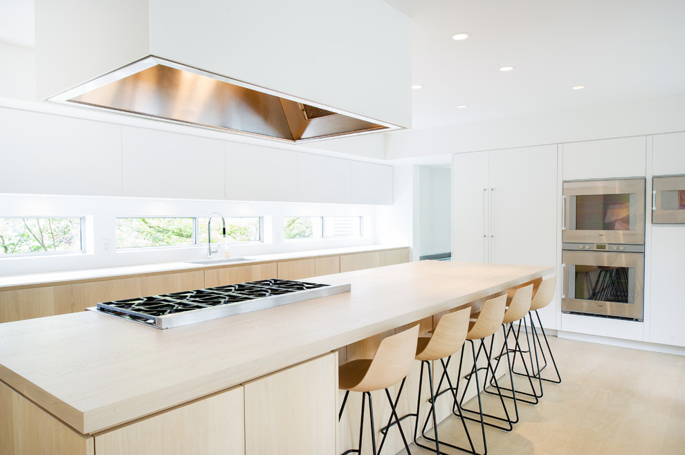Large midcentury open plan kitchen in Vancouver with an undermount sink, flat-panel cabinets, white cabinets, wood benchtops, white splashback, stainless steel appliances, light hardwood floors and with island.