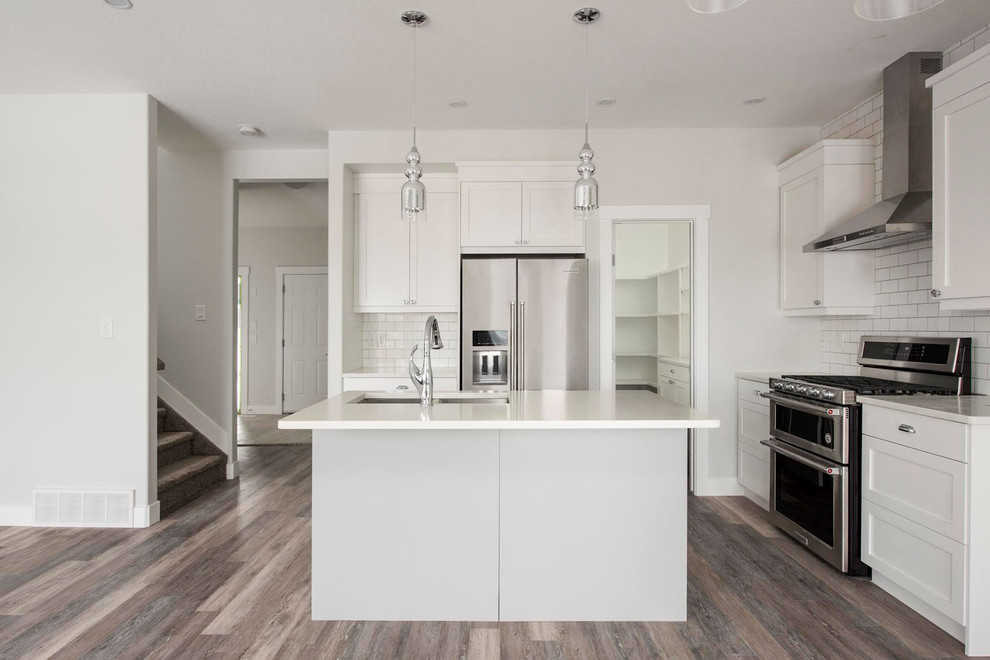 Example of a mid-sized transitional l-shaped medium tone wood floor and brown floor open concept kitchen design in Calgary with an undermount sink, shaker cabinets, white cabinets, quartzite countertops, white backsplash, subway tile backsplash, stainless steel appliances, an island and white countertops