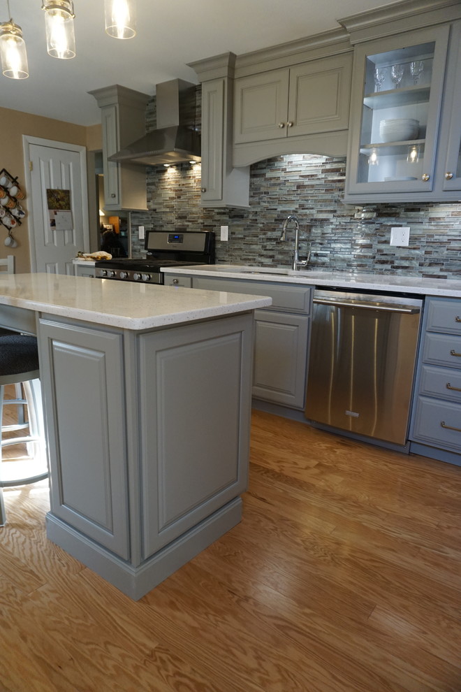 Inspiration for a small timeless l-shaped medium tone wood floor eat-in kitchen remodel in Providence with a triple-bowl sink, raised-panel cabinets, gray cabinets, quartz countertops, gray backsplash, mosaic tile backsplash, stainless steel appliances, an island and white countertops