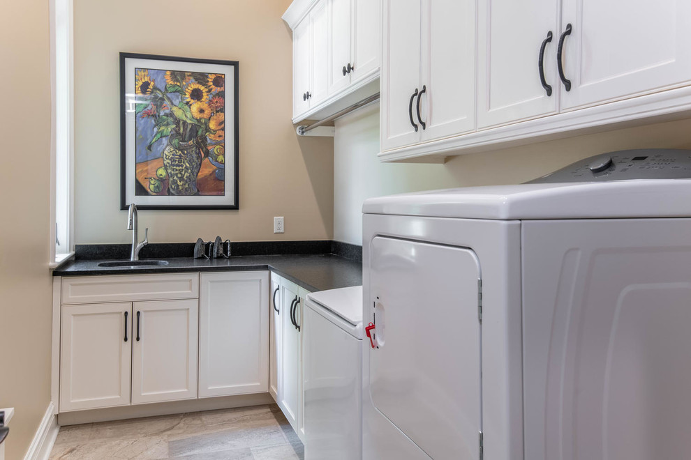 Photo of a mid-sized traditional l-shaped dedicated laundry room in Other with an undermount sink, recessed-panel cabinets, white cabinets, granite benchtops, beige walls, porcelain floors, a side-by-side washer and dryer, beige floor and black benchtop.