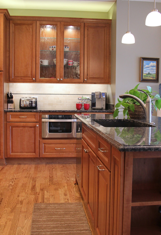 Photo of a large traditional l-shaped open plan kitchen in Milwaukee with an undermount sink, recessed-panel cabinets, brown cabinets, granite benchtops, beige splashback, ceramic splashback, stainless steel appliances, light hardwood floors, multiple islands, brown floor and black benchtop.