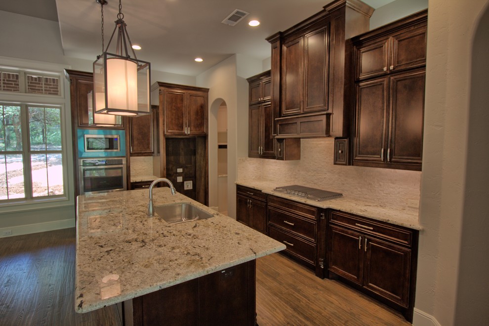Large transitional l-shaped open plan kitchen in Dallas with an undermount sink, raised-panel cabinets, dark wood cabinets, granite benchtops, white splashback, stone tile splashback, stainless steel appliances, medium hardwood floors and with island.