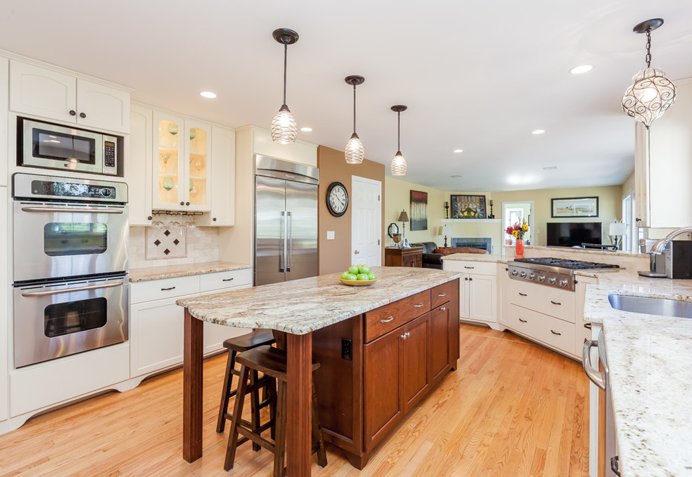 Example of a mid-sized transitional u-shaped light wood floor enclosed kitchen design in Boston with recessed-panel cabinets, granite countertops, beige backsplash, stone tile backsplash, stainless steel appliances, an island, a double-bowl sink and white cabinets