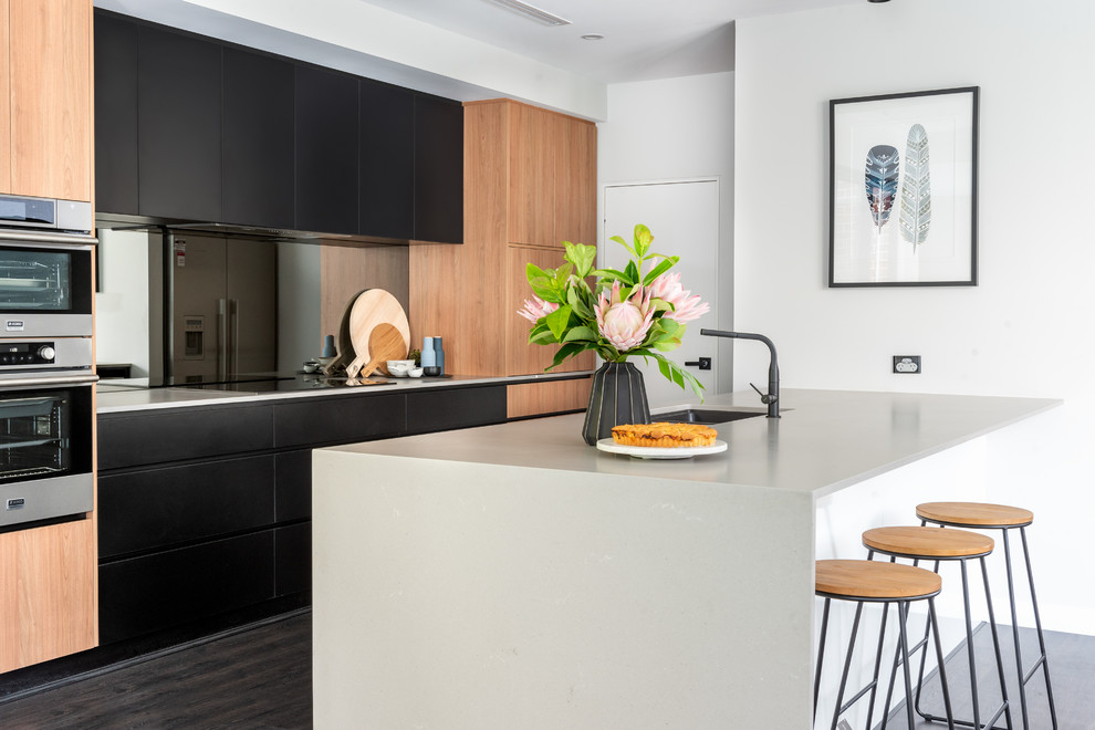 Photo of a contemporary kitchen in Perth with an undermount sink, black cabinets, quartz benchtops, laminate floors, grey benchtop, flat-panel cabinets, grey splashback, glass sheet splashback, stainless steel appliances, a peninsula and black floor.