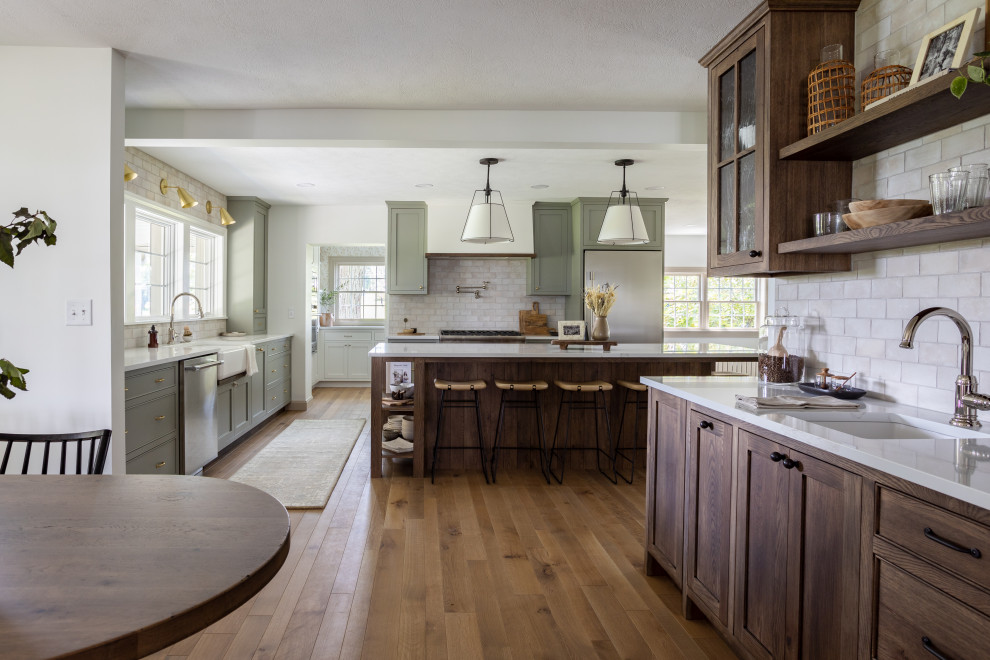 Foto på ett vit kök, med en rustik diskho, skåp i shakerstil, gröna skåp, vitt stänkskydd, stänkskydd i stenkakel, rostfria vitvaror, mellanmörkt trägolv, en köksö och brunt golv