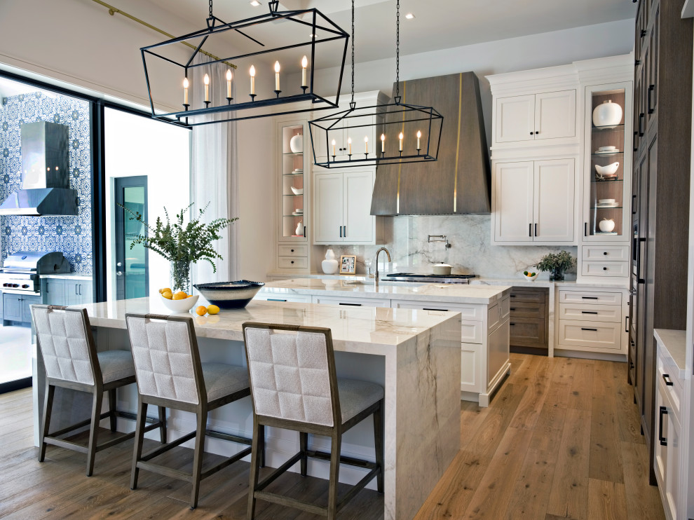 Photo of an expansive transitional l-shaped kitchen pantry in Miami with a farmhouse sink, shaker cabinets, white cabinets, solid surface benchtops, white splashback, porcelain splashback, stainless steel appliances, light hardwood floors, multiple islands, brown floor, white benchtop and recessed.