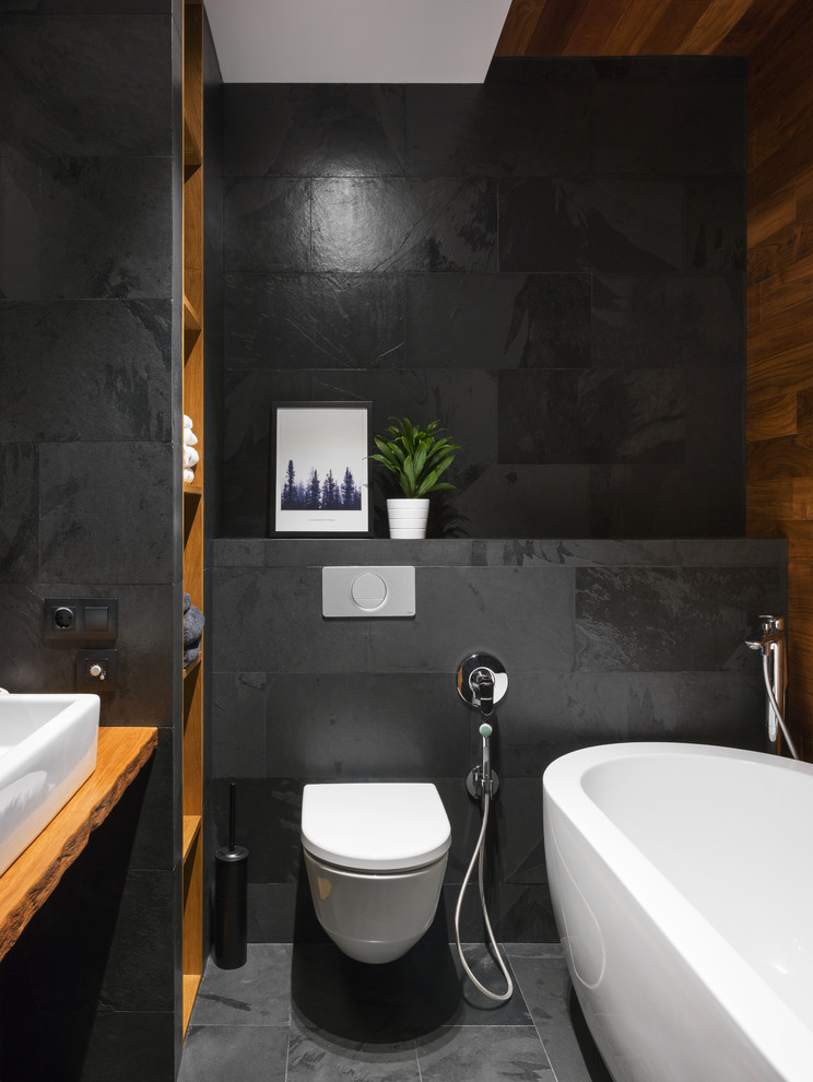 This is an example of a small industrial master bathroom in Moscow with a freestanding tub, a wall-mount toilet, black tile, a vessel sink, wood benchtops, black floor, brown benchtops, open cabinets and black walls.