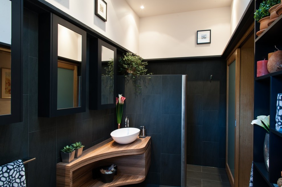 This is an example of a mid-sized modern master bathroom in Sydney with furniture-like cabinets, medium wood cabinets, a freestanding tub, an alcove shower, a one-piece toilet, black tile, porcelain tile, beige walls, porcelain floors, wood benchtops, black floor, an open shower and a vessel sink.