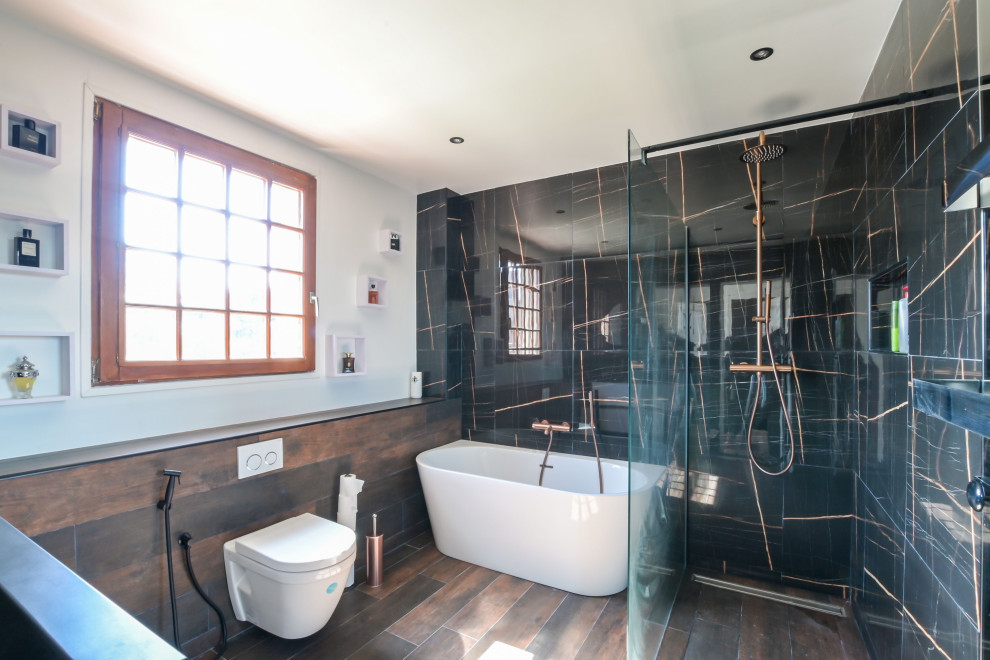 Diseño de cuarto de baño actual con bañera exenta, ducha a ras de suelo, sanitario de pared, baldosas y/o azulejos negros, lavabo de seno grande, suelo marrón y ducha abierta