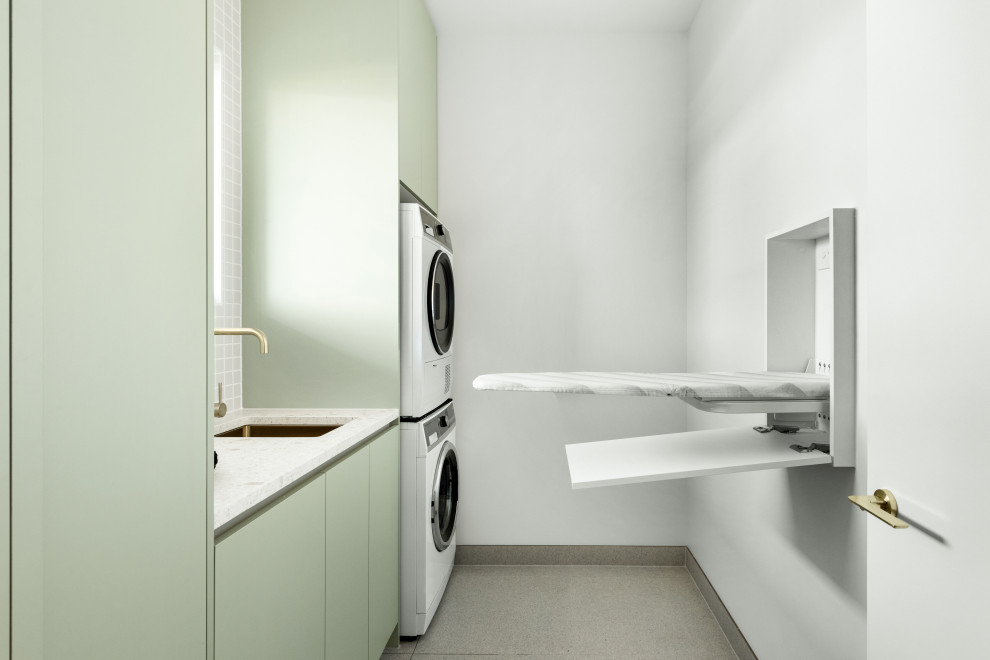 Foto de lavadero lineal actual de tamaño medio con puertas de armario verdes, encimera de terrazo, salpicadero gris, paredes blancas, lavadora y secadora apiladas y encimeras blancas