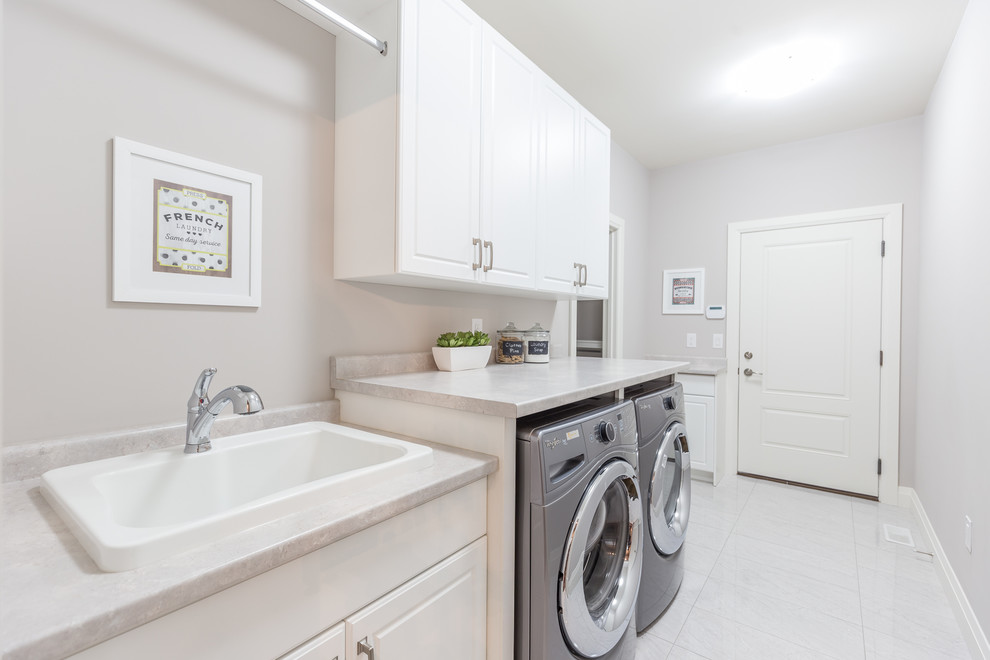 Photo of a large traditional single-wall dedicated laundry room in Toronto with a drop-in sink, raised-panel cabinets, white cabinets, solid surface benchtops, grey walls, travertine floors, a side-by-side washer and dryer and grey floor.