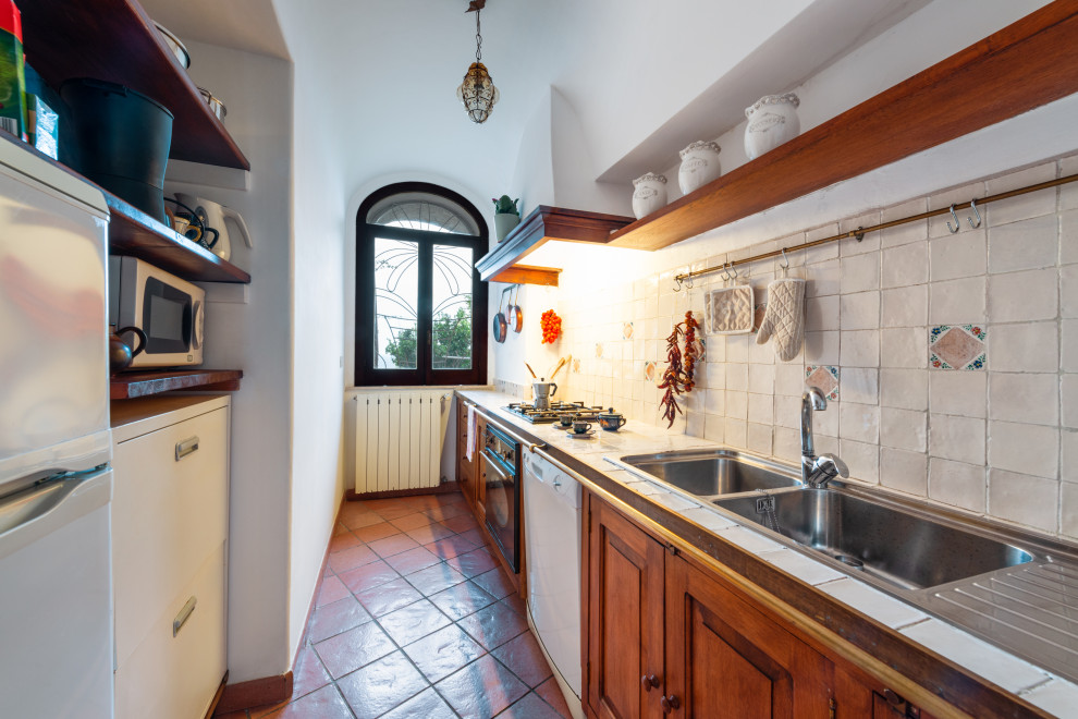 Small mediterranean single-wall separate kitchen in Naples with a double-bowl sink, recessed-panel cabinets, medium wood cabinets, tile benchtops, white splashback, porcelain splashback, white appliances, terra-cotta floors, with island, brown floor, white benchtop and vaulted.