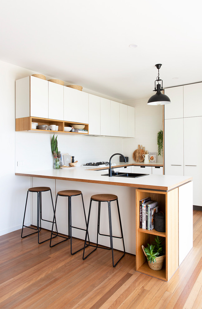 Photo of a beach style u-shaped kitchen in Gold Coast - Tweed with a double-bowl sink, flat-panel cabinets, white cabinets, white splashback, light hardwood floors, a peninsula, beige floor and white benchtop.