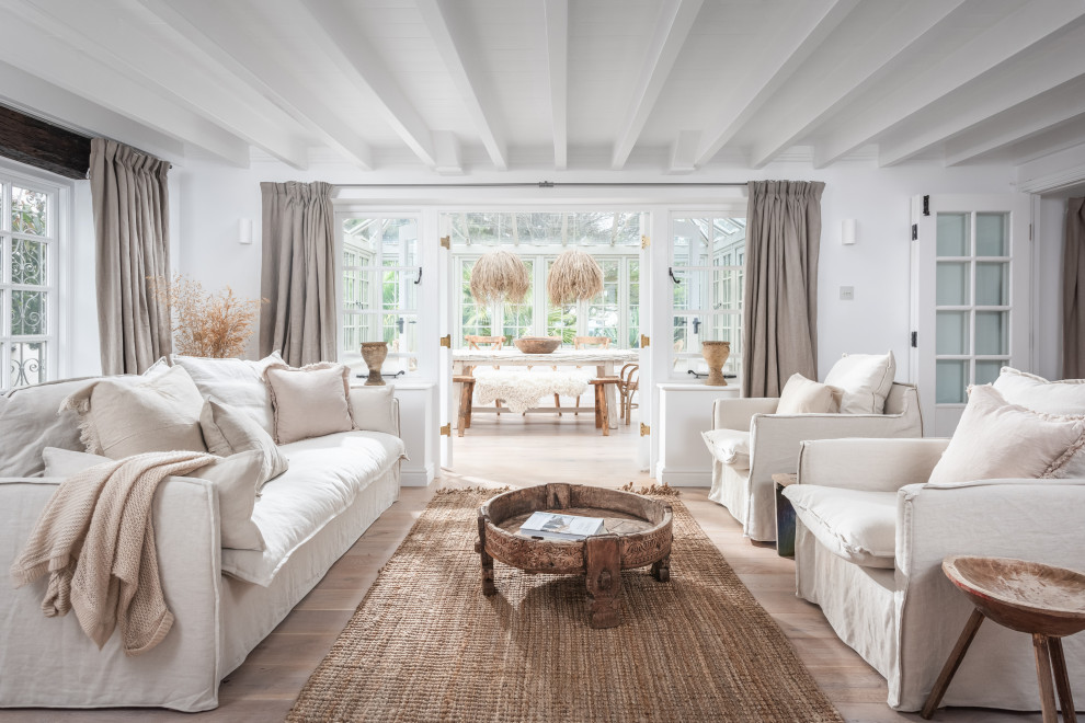 Imagen de salón abierto costero grande con paredes blancas, suelo de madera clara, chimenea de doble cara, televisor retractable y suelo beige