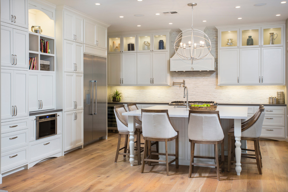 This is an example of a mid-sized transitional l-shaped open plan kitchen in San Diego with stainless steel appliances, light hardwood floors, white cabinets, quartz benchtops, white splashback, stone tile splashback, a double-bowl sink, recessed-panel cabinets and a peninsula.