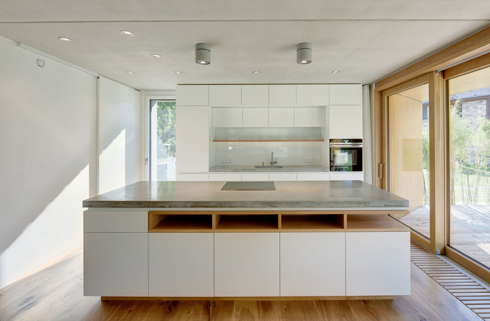 Inspiration for a large contemporary kitchen in Berlin with an integrated sink, flat-panel cabinets, white cabinets, concrete benchtops, white splashback, glass sheet splashback, black appliances, medium hardwood floors and with island.