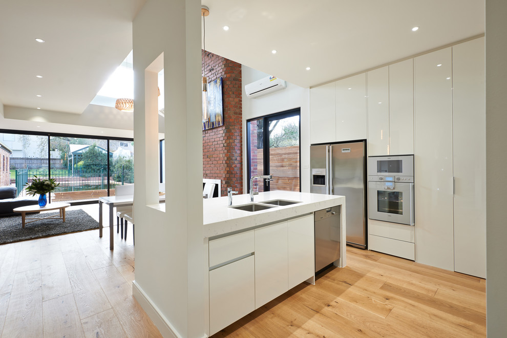 Mid-sized contemporary l-shaped eat-in kitchen in Melbourne with a drop-in sink, flat-panel cabinets, white cabinets, quartz benchtops, metallic splashback, glass sheet splashback, stainless steel appliances, medium hardwood floors and with island.
