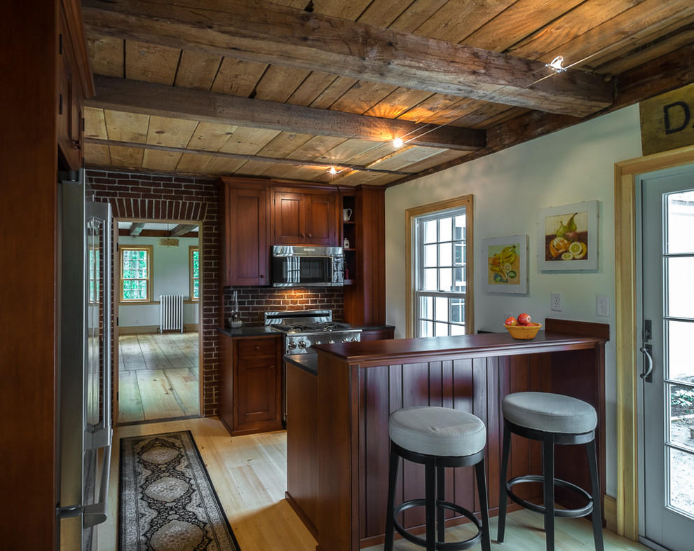 Inspiration for a small country l-shaped separate kitchen in Portland Maine with a farmhouse sink, stainless steel appliances, light hardwood floors, a peninsula, recessed-panel cabinets, dark wood cabinets, soapstone benchtops and red splashback.