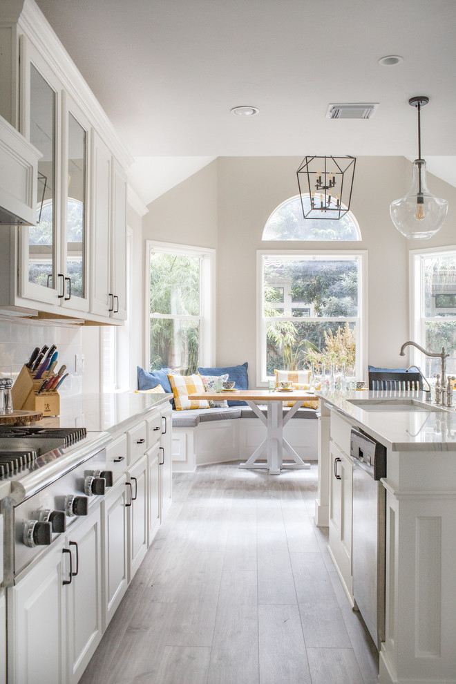Inspiration for a traditional galley eat-in kitchen in Houston with an undermount sink, raised-panel cabinets, white cabinets, white splashback, subway tile splashback, stainless steel appliances, light hardwood floors, with island, grey floor and white benchtop.