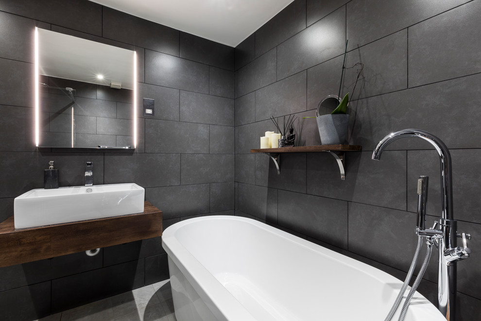 Photo of a large modern bathroom in London with raised-panel cabinets, dark wood cabinets, a freestanding tub, a shower/bathtub combo, a one-piece toilet, gray tile, cement tile, grey walls, ceramic floors, a pedestal sink and wood benchtops.