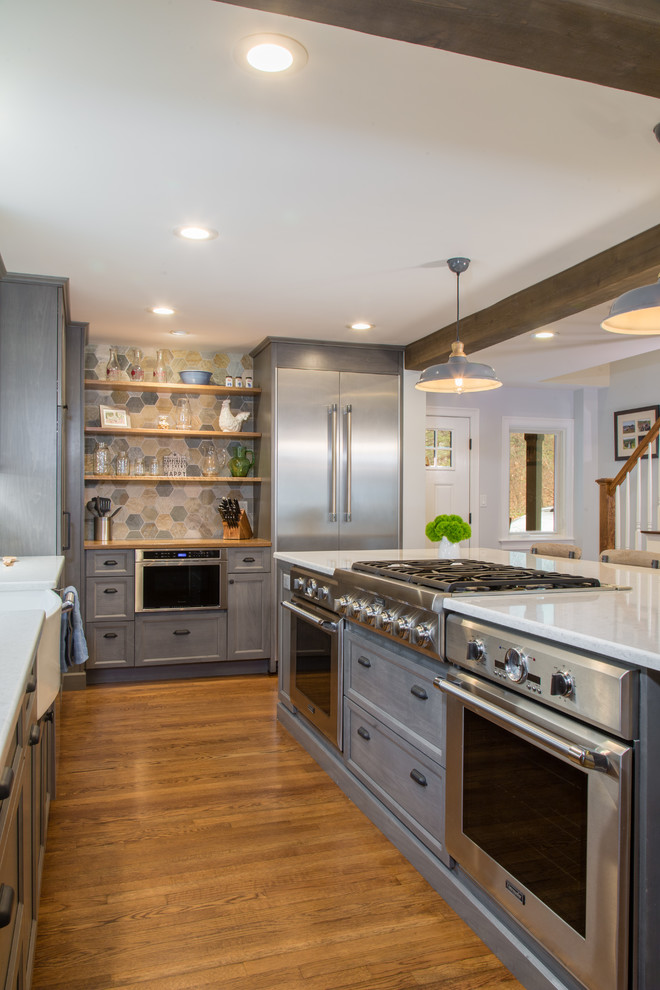 Country l-shaped kitchen in Boston with a farmhouse sink, shaker cabinets, grey cabinets, stainless steel appliances, medium hardwood floors, with island, brown floor and white benchtop.