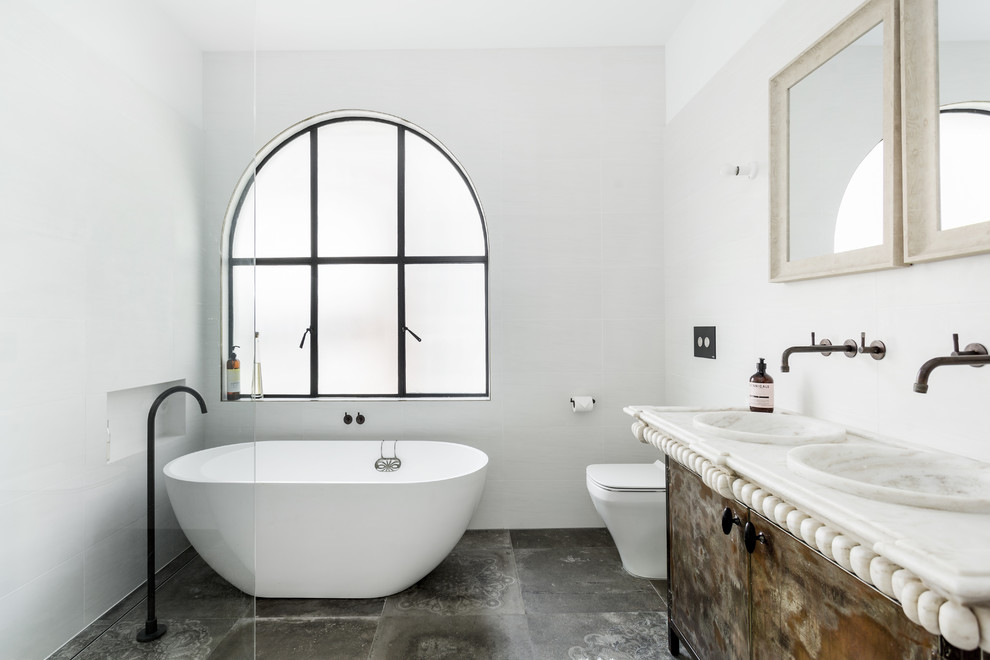 Design ideas for a mid-sized mediterranean master bathroom in Sunshine Coast with flat-panel cabinets, distressed cabinets, a freestanding tub, a one-piece toilet, grey walls, an integrated sink, marble benchtops, grey floor, beige benchtops and ceramic floors.