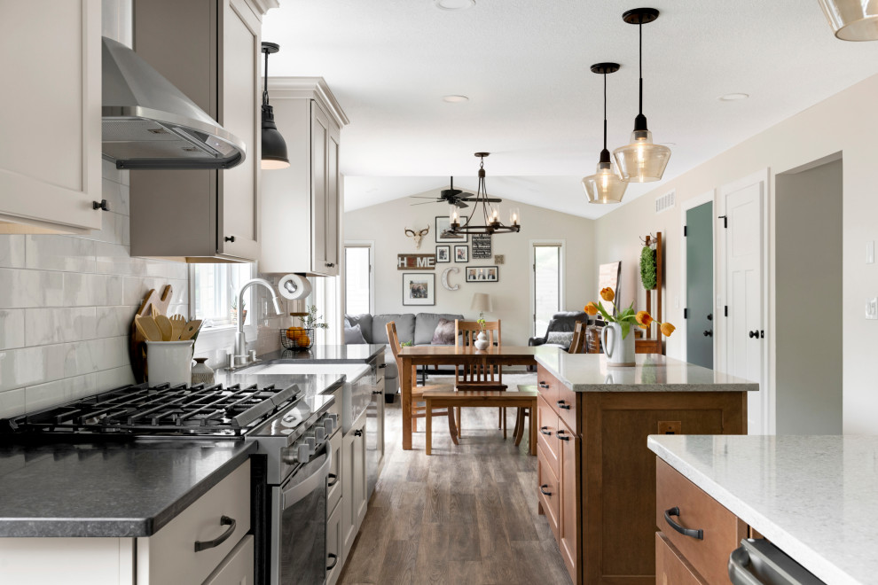 Photo of a large transitional single-wall eat-in kitchen in Minneapolis with a farmhouse sink, shaker cabinets, grey cabinets, quartz benchtops, white splashback, ceramic splashback, stainless steel appliances, vinyl floors, multiple islands, brown floor and black benchtop.
