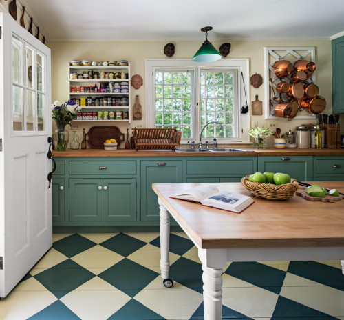 Rolling mobile island with butcher-block top on casters adds extra counter space beside green shaker kitchen cabinet, open spice shelves, and copper pot rack in a bright cottage kitchen