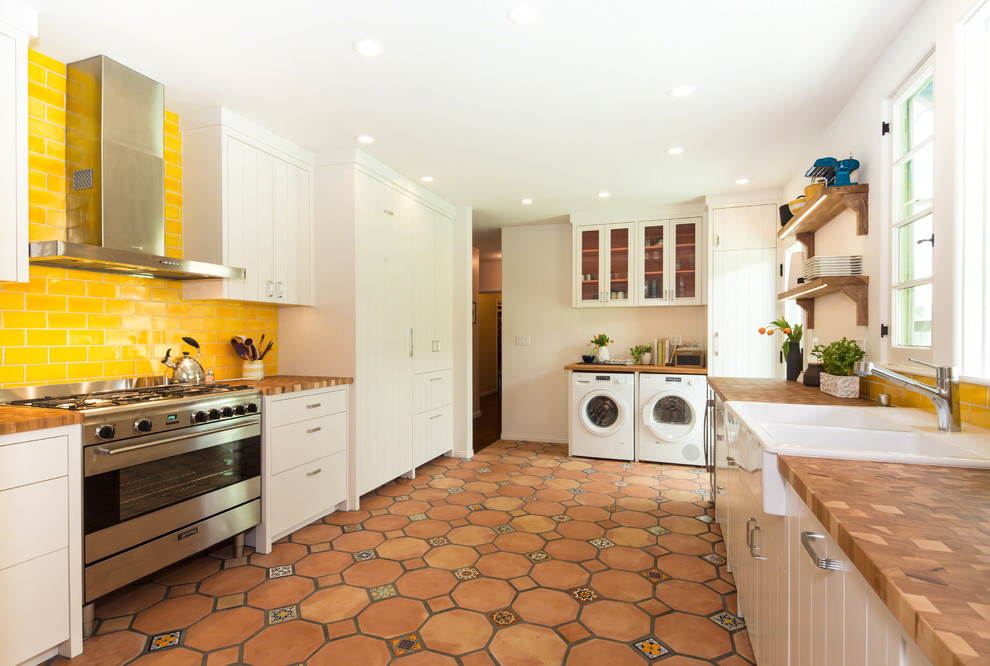 Design ideas for a transitional u-shaped kitchen in Los Angeles with a farmhouse sink, flat-panel cabinets, white cabinets, yellow splashback, subway tile splashback, stainless steel appliances, terra-cotta floors and no island.