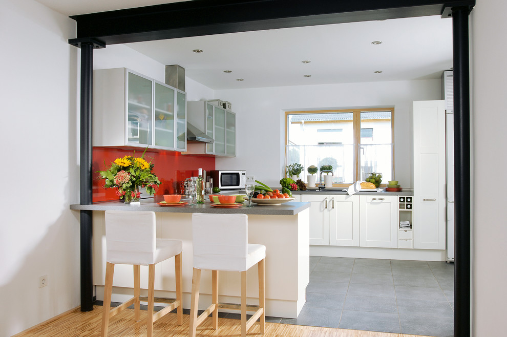 This is an example of a mid-sized contemporary u-shaped open plan kitchen in Cologne with recessed-panel cabinets, white cabinets, red splashback, glass sheet splashback, a peninsula, ceramic floors, grey floor and grey benchtop.