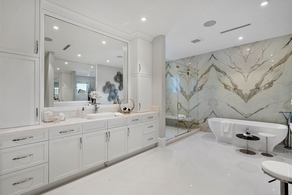 Идея дизайна: огромная главная ванная комната в стиле неоклассика (современная классика) с белыми фасадами, отдельно стоящей ванной, душем в нише, унитазом-моноблоком, плиткой из листового камня, бежевыми стенами, мраморным полом, накладной раковиной, мраморной столешницей, бежевым полом, душем с распашными дверями, белой столешницей, тумбой под одну раковину и встроенной тумбой