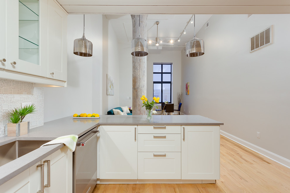 This is an example of an industrial open plan kitchen in New York with an undermount sink, shaker cabinets, white cabinets, stainless steel appliances, light hardwood floors, a peninsula and beige floor.