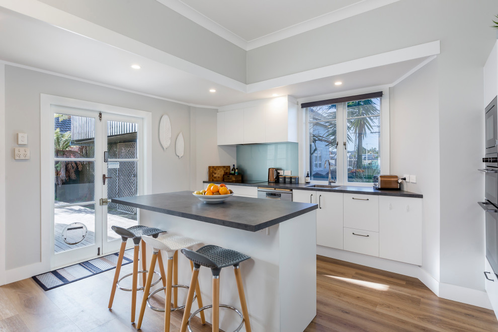 Photo of a mid-sized modern l-shaped eat-in kitchen in Auckland with a drop-in sink, flat-panel cabinets, white cabinets, laminate benchtops, glass sheet splashback, stainless steel appliances, with island, black benchtop and blue splashback.