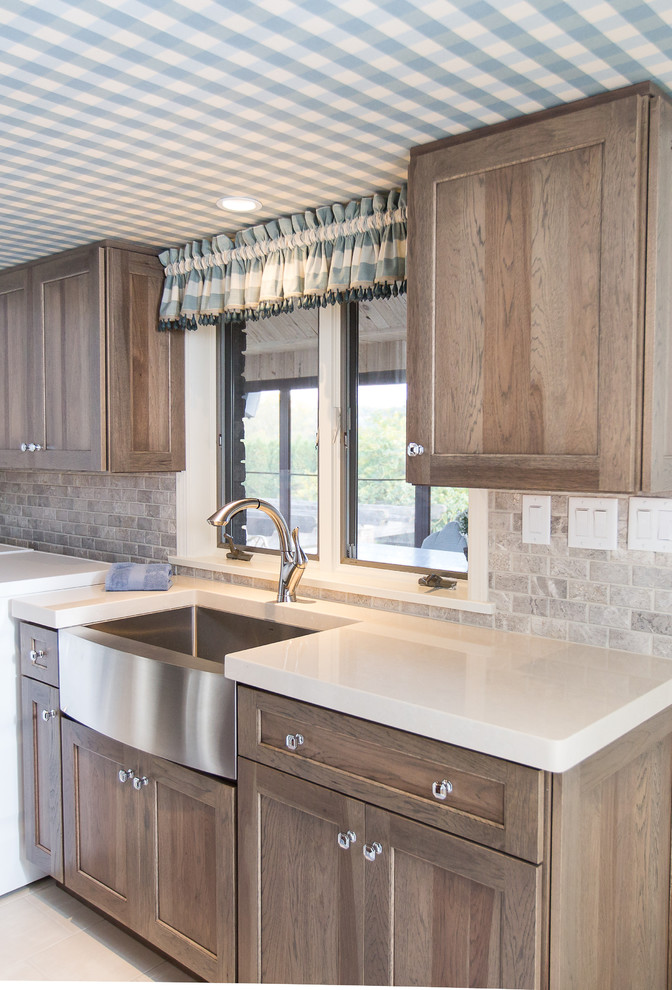 Photo of a mid-sized country galley dedicated laundry room in Other with shaker cabinets, solid surface benchtops, grey splashback, subway tile splashback, a farmhouse sink, medium wood cabinets, ceramic floors and a side-by-side washer and dryer.