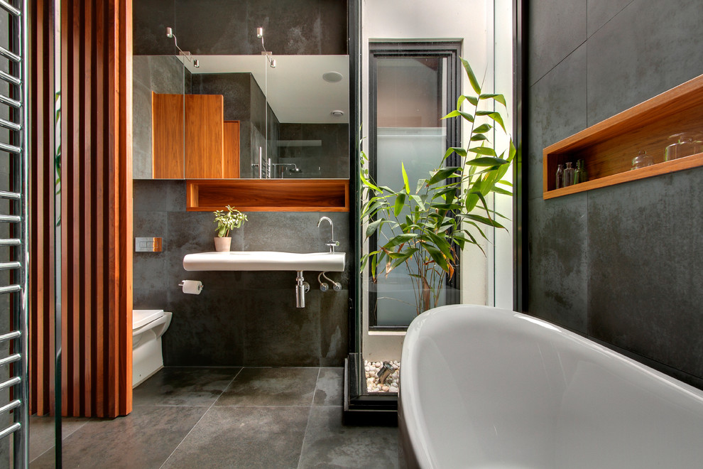 Contemporary bathroom in Melbourne with a wall-mount sink, medium wood cabinets, a freestanding tub, gray tile, grey walls, a one-piece toilet and a niche.