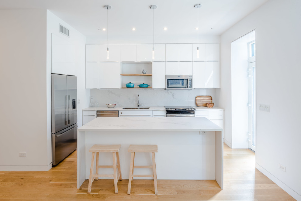 Mid-sized modern l-shaped eat-in kitchen in New York with an undermount sink, flat-panel cabinets, white cabinets, glass benchtops, white splashback, stone slab splashback, stainless steel appliances, light hardwood floors, with island and white benchtop.