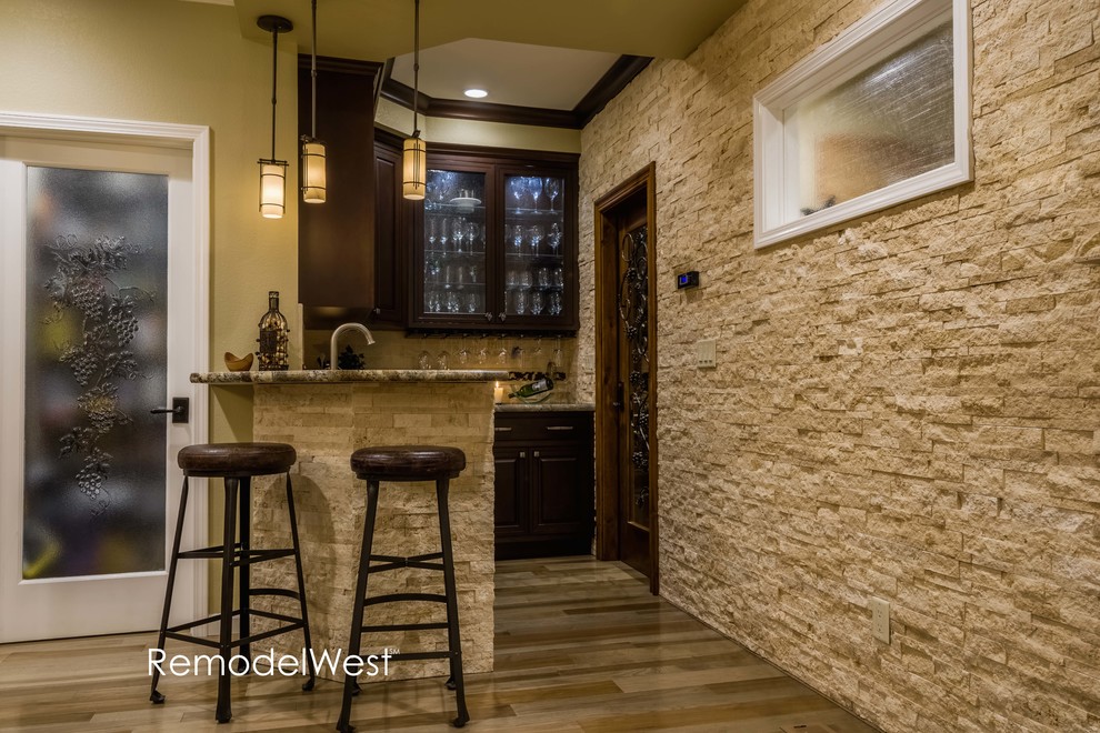 This is an example of a small transitional l-shaped seated home bar in San Francisco with an undermount sink, raised-panel cabinets, dark wood cabinets, granite benchtops, beige splashback, stone tile splashback and light hardwood floors.